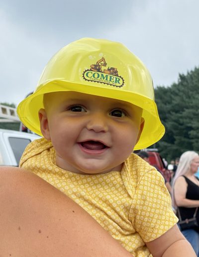A smiling baby with a Comer Construction hard hat.