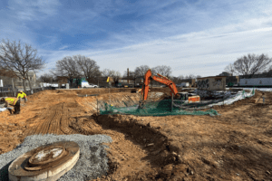 Hitachi excavating mid-site, manhole lid in foreground gravel
