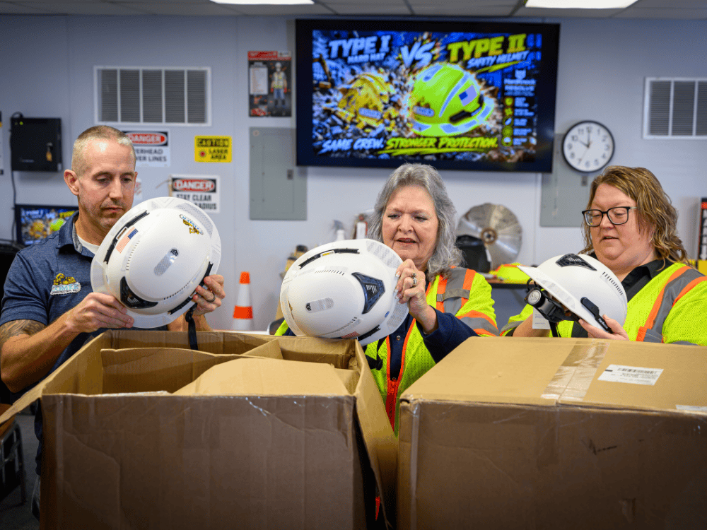 Comer Construction team members unpack new Type II safety helmets in front of a screen comparing Type I hard hats and Type II helmets.